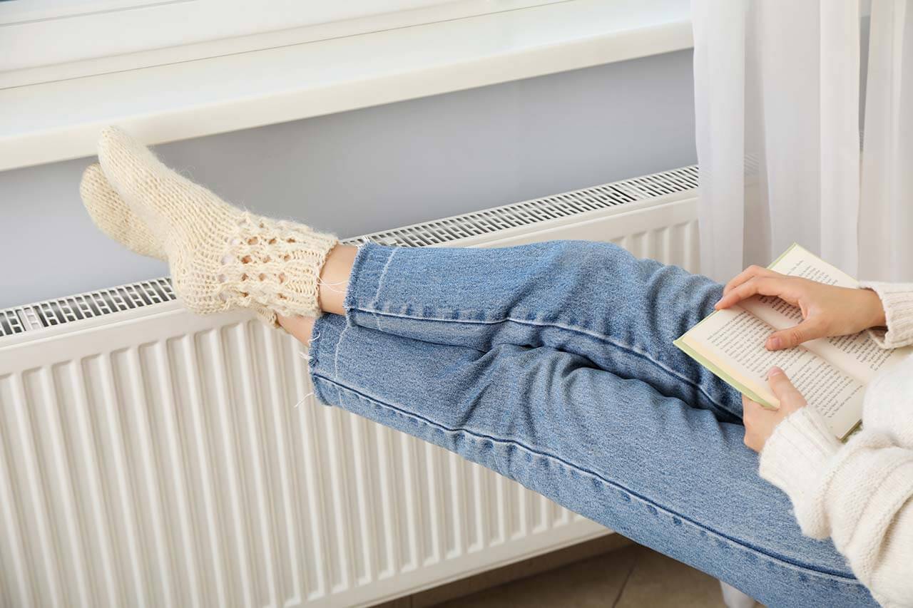 Person warming up by a radiator in a cozy room, showcasing the need for reliable baseboard heating.